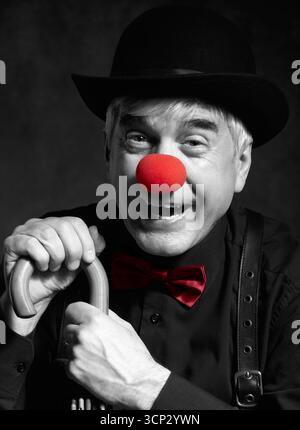 Selektives Farbporträt eines lachenden Clowns am Griff eines Schirms. Ein älterer Mann mit roter Clownnnase und Bowler-Hut auf dem Kopf. ich war angezogen Stockfoto
