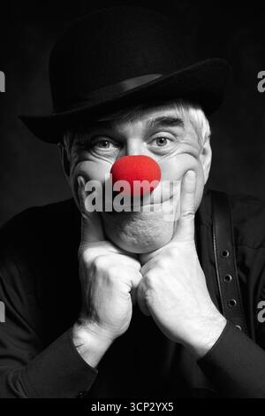 Dramatisch selektives Farbporträt eines traurigen Clowns vor dunklem Hintergrund. Ein älterer Mann mit roter Clownnnase und Bowler-Hut auf dem Kopf. Gekleidet in Blac Stockfoto