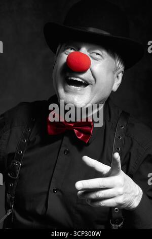 Selektives Farbporträt eines lachenden Clowns vor dunklem Hintergrund. Ein älterer Mann mit roter Clownnnase und Bowler-Hut auf dem Kopf. In schwarzem Shir gekleidet Stockfoto