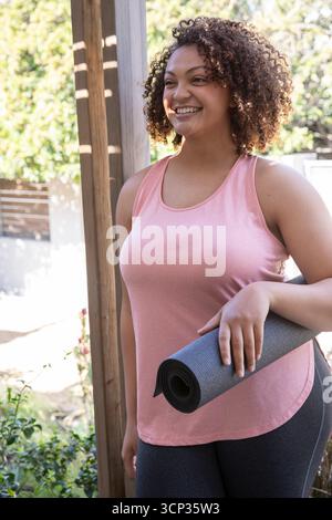 Afroamerikanerin, die rosa Posen auf der Terrasse trägt, Yoga-Matte gegen Post hält, Kopierraum Stockfoto