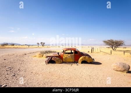 Auf dem Autofriedhof von Solitaire in Namibia befinden sich rostige Oldtimer in der Nähe kleiner Wüstengebäude, die vor Dünen und Bergen liegen und den Charme der Wüste einfangen Stockfoto