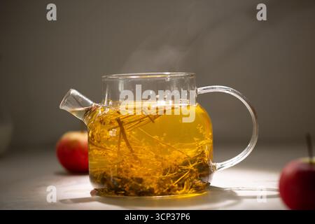 Eine Teekanne aus klarem Glas tränkt Kräuter in heißes Wasser und gibt einen leichten Dampf ab. Zwei rote Äpfel stehen auf der Theke neben der Teekanne, tagsüber drinnen. Stockfoto