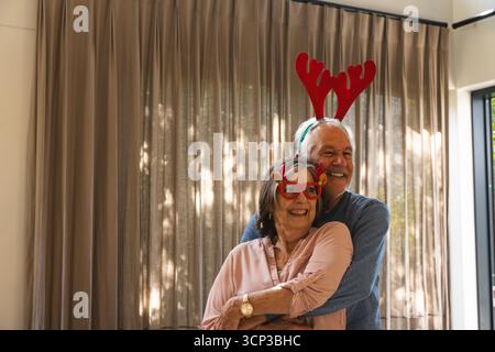 Seniorenpaar, das sich im Wohnzimmer umgibt, während es eine rote Geweihbrille und ein Stirnband am Fenster trägt Stockfoto