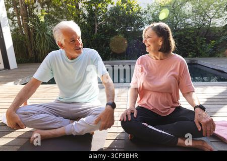 Ein älteres Paar sitzt auf Yogamatten auf der Hinterhofterrasse neben dem Pool und trägt T-Shirts und Fitness-Tracker Stockfoto