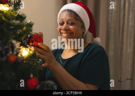 Ältere Afroamerikanerin mit Weihnachtsmann-Hut, die rote Kugel am Weihnachtsbaum im Wohnzimmer hängt Stockfoto