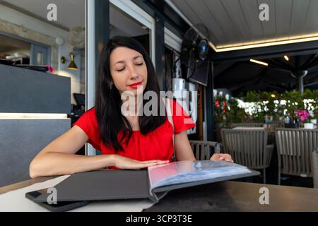 Frau liest Menü am Outdoor Restaurant Tisch. Eine elegante Frau in rotem Kleid stöbert in einem modernen Restaurant im Freien, umgeben von einem Menü Stockfoto