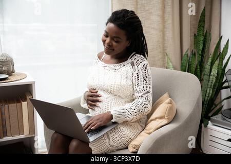 Eine schwangere Frau sitzt bequem zu Hause und lächelt, während sie ihren Laptop benutzt. Stockfoto