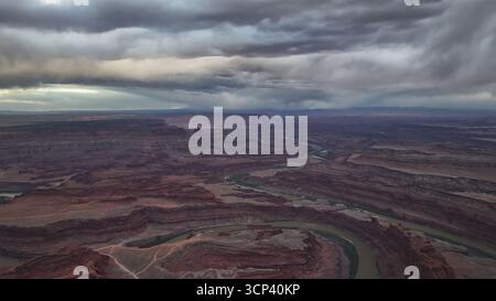 Aus der Vogelperspektive auf den Colorado River, der sich durch die dramatischen roten Felsschluchten unter einem stürmischen Himmel schlängelt, ein Wandteppich aus kontrastierenden Texturen und Tönen, Moab, Utah, USA. Stockfoto