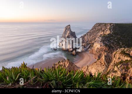 Der Blick auf die Golden Hour zeigt die zerklüfteten Klippen und den abgeschiedenen Strand, wo die Wellen des Atlantiks sanft abstürzen und eine ruhige Küstenszene schaffen, Praia da Ursa, Lissabon, Portugal. Stockfoto