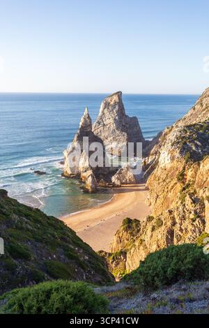 Blick auf zerklüftete Klippen treffen auf den weichen Sandstrand, wo schäumende Wellen in goldenem Sonnenlicht stürzen. Praia da Ursa, Lissabon, Portugal, ist eine Oase natürlicher Schönheit an der Küste. Stockfoto