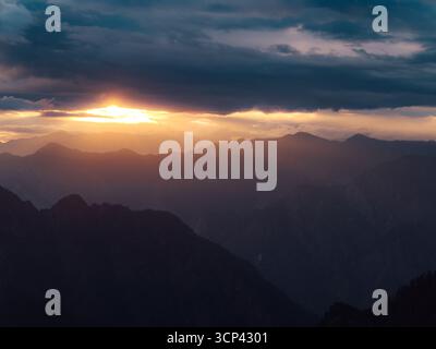 Aus der Vogelperspektive blickt die Sonne durch dunkle, schwere Wolken und strahlt ein warmes, ätherisches Licht über die zerklüftete Bergkette in Sosan, Himachal Pradesh, Indien. Stockfoto