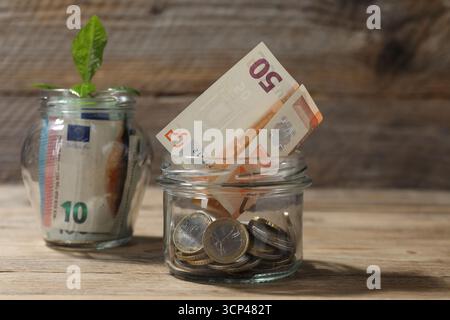 Glasgläser mit Münzen, Sprossen und Banknoten auf Holztisch, Nahaufnahme Stockfoto