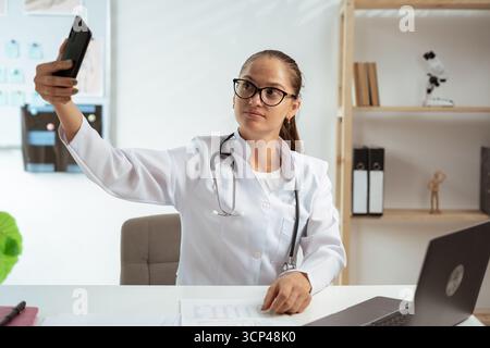 Ärztin beim Selfie-Foto in der Krankenhauspraxis. Die Ärztin führt Videoanrufe von ihrem Arbeitsplatz aus. Kopierbereich Stockfoto