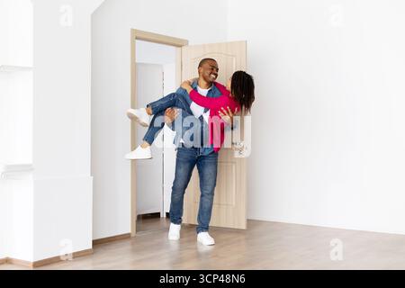 Glücklicher schwarzer Mann, der einen lächelnden Partner in den Armen trägt, während er ein neues Zuhause betritt Stockfoto
