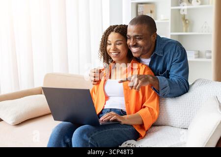 Lächelndes afroamerikanisches Paar sitzt auf Sofa mit Laptop und genießt die gemeinsame Zeit Stockfoto