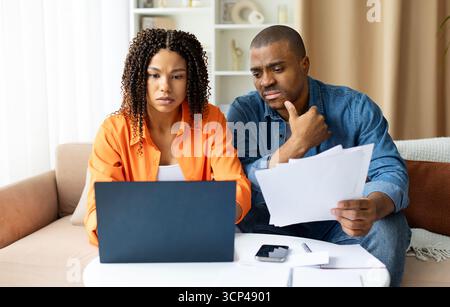 Afroamerikanisches Paar, das mit Papieren am Laptop arbeitet und zu Hause besorgt aussieht Stockfoto