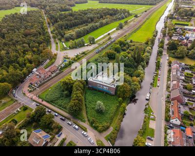 Aus der Vogelperspektive eines halbländlichen Gebiets mit Eisenbahn, Bahnhof, Kanal mit Booten, solargedecktem Gebäude, roten Häusern, Wald und Felder werden gemischt Stockfoto