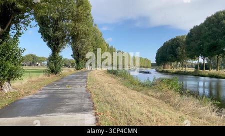 Tournai Belgien September 2025. Bäume säumen die Schelde mit einem Radweg entlang. Ein Frachtschiff fährt Richtung Tournai Schelde, Binnenland, Wasserstraßen, Schifffahrt, Infrastruktur, Stockfoto