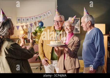 Ältere Frau hält Geburtstagskuchen, während Freunde lächeln und posieren Stockfoto