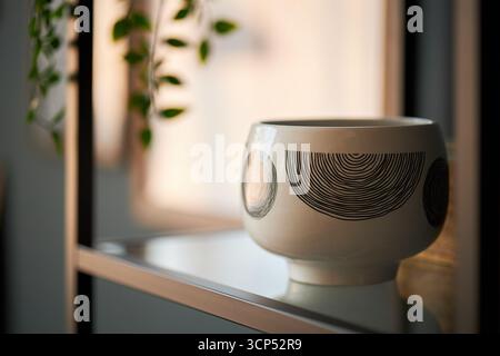 Keramikschale mit abstrakten Kreismustern, die auf einem Glasregal in der Nähe des Fensters steht, weiches natürliches Licht hebt glatte Oberfläche hervor, grüne Pflanzenblätter hängen im Hintergrund Stockfoto