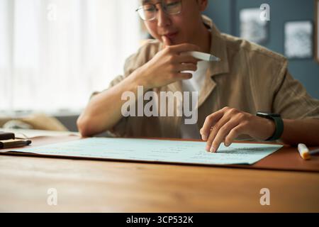 Asiatischer junger Erwachsener, der am Schreibtisch sitzt, Dokumente analysiert und Stift hält, Brille trägt und sich auf Papierkram konzentriert, partielles Gesicht sichtbar, natürliche Inneneinrichtung Stockfoto