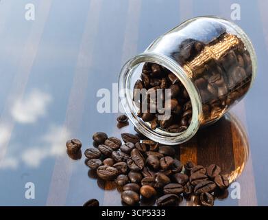 Kaffeebohnen, die aus dem Glasbehälter verschüttet werden, geröstete Kaffeebohnen auf reflektierender Oberfläche Stockfoto