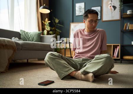 Asiatischer junger erwachsener Mann, der auf Teppichboden sitzt und vom Smartphone in einem modernen Wohnzimmer wegschaut, nachdenklich und entspannt wirkt, natürliches Licht aus dem Fenster kommt Stockfoto