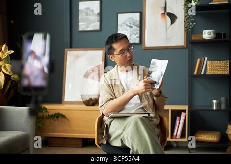 Asiatischer junger erwachsener Mann sitzt und erklärt das Foto, während er auf das gedruckte Bild drückt, das Notebook auf dem Schoß hält, Smartphone auf einem Stativ, das Video in einem modernen Raum aufnimmt Stockfoto