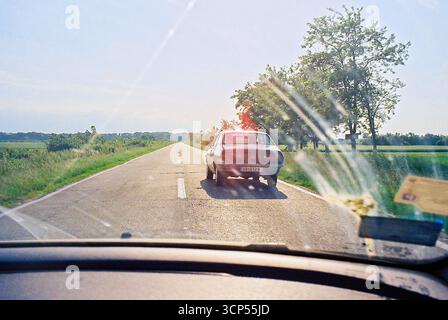 Analoges Foto vom Beifahrersitz, das einen alten Ford zeigt, der voraus fährt. Roadtrip-Szene mit Retro-Stimmung und Bewegung. Stockfoto