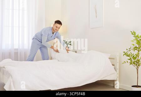 Junge Männerschwester in blauer Uniform, die weißes Bett in der modernen medizinischen Klinik macht Stockfoto