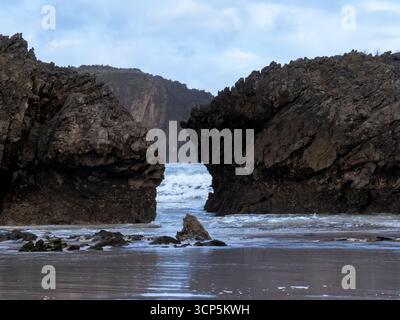 Wellen, die sanft in einen Strand zwischen großen Felsformationen unter einem bewölkten Himmel eindringen Stockfoto