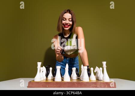 Attraktive Frau, die sich in einem Schachspiel engagiert, zeigt Strategie und Fokus auf einem soliden khakigrünen Hintergrund Stockfoto