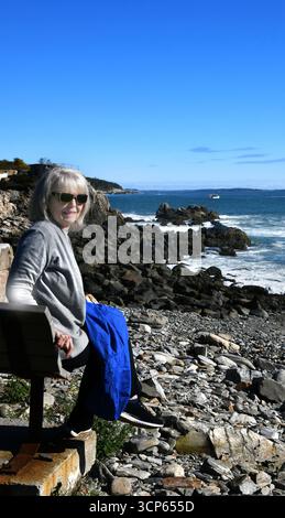 Seniorin sitzt auf einer Bank in Fort Williams Park, Portland, Maine. Sie trägt eine Sonnenbrille und eine Jacke. Sie schaut in die Kamera Stockfoto