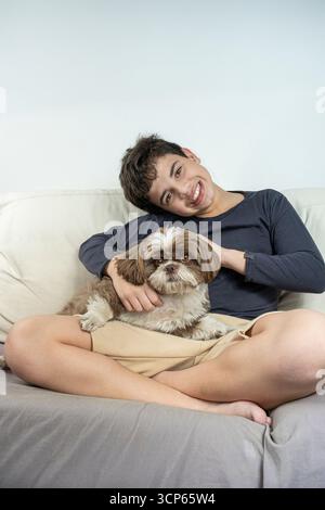 Lächelnder 12-jähriger brasilianischer Teenager auf dem Sofa neben seinem Hund vertikal. Stockfoto