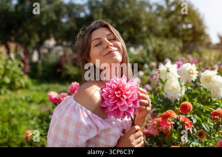 Porträt einer attraktiven Gärtnerin, die im Sommergarten Dahlien auf einem großen Teller hält. Glücklicher junger Bauer, der rosa Blüten auf dem Feld pflückt. Stockfoto