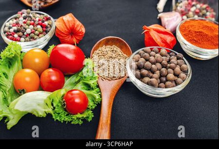 Hölzerne Löffel von Kümmelsamen zira Nahaufnahme unter verschiedenen Arten von Paprika in Schüsseln, geräucherter Paprika und verschiedenen Gemüse auf schwarzem Hintergrund. Culina Stockfoto