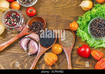 Holzlöffel mit Gewürzen von Kümmel, schwarzem Sesam und Nelken. Verschiedene Arten von Pfefferkörnern in Schüsseln. Salat, Kirschtomaten, Physalis, Knoblauch. Holz Stockfoto