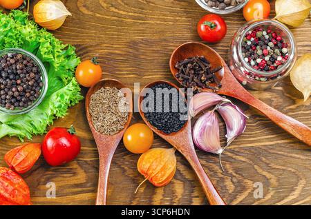Holzlöffel mit Gewürzen von Kümmel, schwarzem Sesam und Nelken. Verschiedene Arten von Pfefferkörnern in Schüsseln. Salat, Kirschtomaten, Physalis. Holzoberfläche Stockfoto