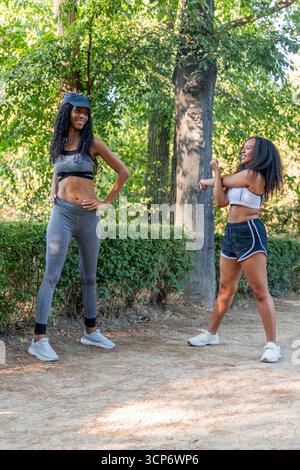 Vertikal zwei lächelnde lateinamerikanische Frauen in lebendiger Sportswear, die sich in einem Park ausstrecken und sich auf ein Lauftraining mit einer hellen und sonnigen Summe vorbereiten Stockfoto
