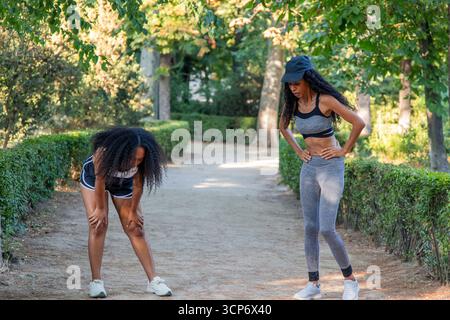 Zwei müde lateinamerikanische Sportlerinnen erholten sich nach einem anspruchsvollen Lauf im Park, schnappen sich den Atem, während sie ihre Beine Strecken und die Wärme von genießen Stockfoto