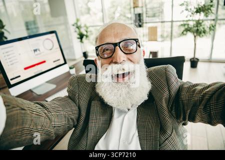 Geschäftsmann in stilvoller Formalwear, der ein fröhliches Selfie in einer modernen Büroumgebung mit Tageslicht macht Stockfoto