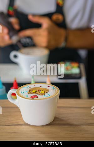 Niedliche Latte Art in Form einer Zeichentrickkatze mit bunten Elementen, serviert in einer weißen Tasse auf Holztisch. Konzept von Latte Art, Kaffee Kreativität und Spaß Café Stockfoto