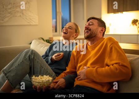 Lachende, fröhliche Paare essen Popcorn und schauen zusammen auf dem Sofa zu Hause fern, Familienspaß Stockfoto