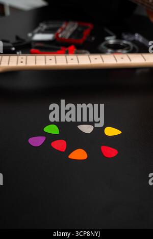 Eine Reihe von bunten Gitarrenpicks liegt auf einer schwarzen Oberfläche mit Gitarren und Reparaturwerkzeugen. Mehrere Gitarren-Picks in hellen Farben auf dem Tisch. Gitarrenauswahl Stockfoto