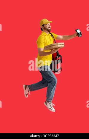 Männlicher Kurier mit Pizzakästen und Zahlungsterminal, der auf rotem Hintergrund springt Stockfoto