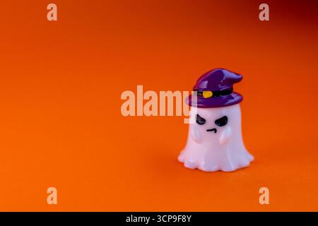 Der Geist trägt einen lila Hexenhut mit bösem Look auf orangefarbenem Hintergrund unter dramatischem Rampenlicht. Verspielte Halloween-Dekoration, perfekt für die Saison Stockfoto