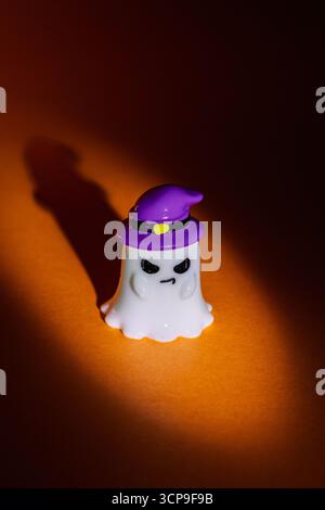 Der Geist trägt einen lila Hexenhut mit bösem Look auf orangefarbenem Hintergrund unter dramatischem Rampenlicht. Verspielte Halloween-Dekoration, perfekt für die Saison Stockfoto