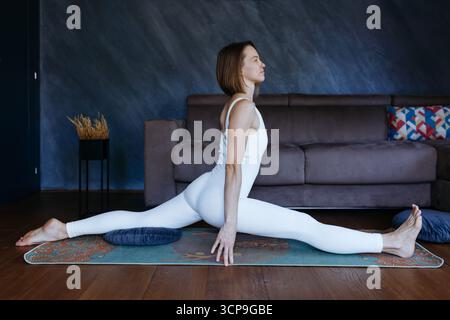 Hellhaarige Frau, die zu Hause Yoga macht, sitzt in Front Splits Pose Stockfoto