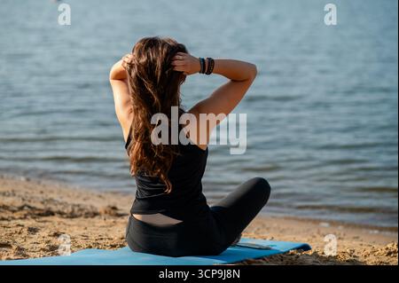 Rückansicht einer Frau in Sportbekleidung, die am Ufer eines Sees sitzt, in die Ferne blickt, ruhige Natur und friedliche Atmosphäre im Freien genießt Stockfoto