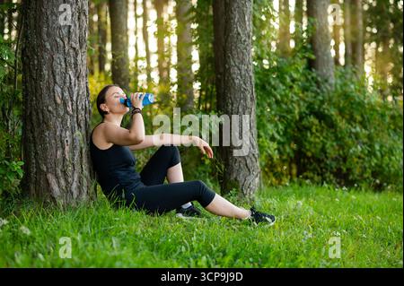 Junge Frau in Sportkleidung trinkt Wasser aus einer Sportflasche, ruht nach dem Joggen im Wald an einem Baum, genießt Feuchtigkeit und die Ruhe der Natur atmos Stockfoto